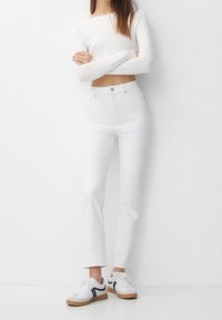 Žena stojící s překříženýma rukama, oblečená v bílém dlouhém crop topu, bílých džínách s vysokým pasem a bílých teniskách.