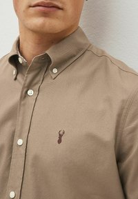 Camicia button-up marrone chiaro realizzata in tessuto liscio; presenta un piccolo emblema di cervo marrone scuro sul petto e colletto classico.