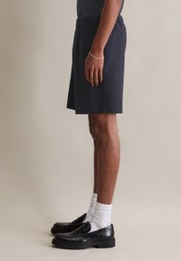 Marineblauwe shorts van gestructureerde stof, met een elastische tailleband. Gemaakt met zwarte instappers en witte enkelsokken. Zijprofielweergave.