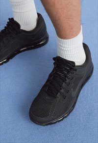 Calçados atléticos pretos com tecido em malha, sistema de atacadores e amortecimento de ar visível. Usados com meias brancas até ao tornozelo em uma superfície azul.