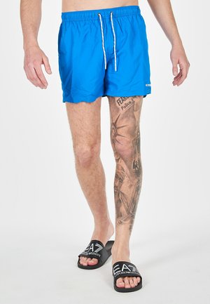 Pantaloni da bagno blu brillante con vita elastica e coulisse, abbinati a ciabatte nere con un logo bianco.