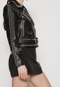 Veste en cuir noir de motard ornée de clous en argent, fermeture éclair asymétrique, poche latérale et taille ceinturée. Fini lisse avec des accents métalliques.