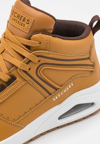 Brown synthetic high-top sneaker with textured accents, orange laces, a cushioned collar, and white midsole featuring air cushion detailing.
