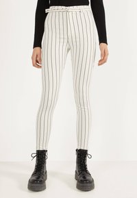 Vita höga byxor med svarta vertikala ränder, med ett bälte och en slim fit-design. Kombinerat med svarta snörade kängor.