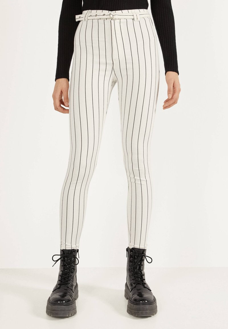 Vita höga byxor med svarta vertikala ränder, med ett bälte och en slim fit-design. Kombinerat med svarta snörade kängor.