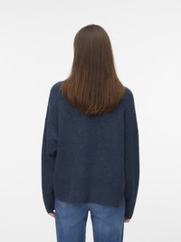 Person mit langen braunen Haaren, der einen dunkelblauen Pullover und blaue Jeans trägt und vor einem schlichten weißen Hintergrund steht.