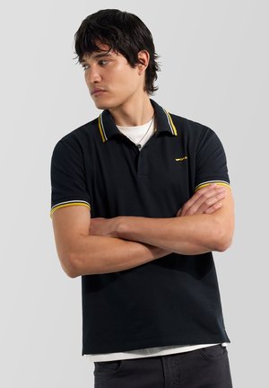 Giovane uomo con le braccia incrociate che indossa una polo nera con colletto e polsini a righe gialle, guarda verso sinistra su sfondo semplice.