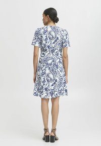 Kratka obleka s kratkimi rokavi, ki prikazuje modro in belo paisley vzorčenje, razširjen rob, oprijet pas in teksturirano tkanino. Nosi se z rjavimi čevlji na blok peto.