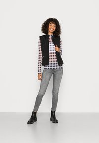 Colete acolchoado preto sobre uma blusa de mangas longas com padrão xadrez multicolorido, combinado com jeans skinny cinzentos e botas pretas de tornozelo.