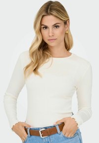 Blonde vrouw met golvend haar in een witte lange mouwen top, blauwe jeans en een bruine riem, met haar handen in de zakken tegen een effen achtergrond.