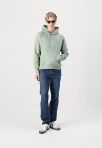 Felpa verde con coulisse e tasca frontale, abbinata a jeans blu. Il modello indossa occhiali da sole e scarpe sportive con strisce bianco e nere.