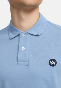 Lyseblå polo skjorte med teksturert stoff, to knapper ved kragen og en svart sirkulær logo med et stilisert hvitt emblem.