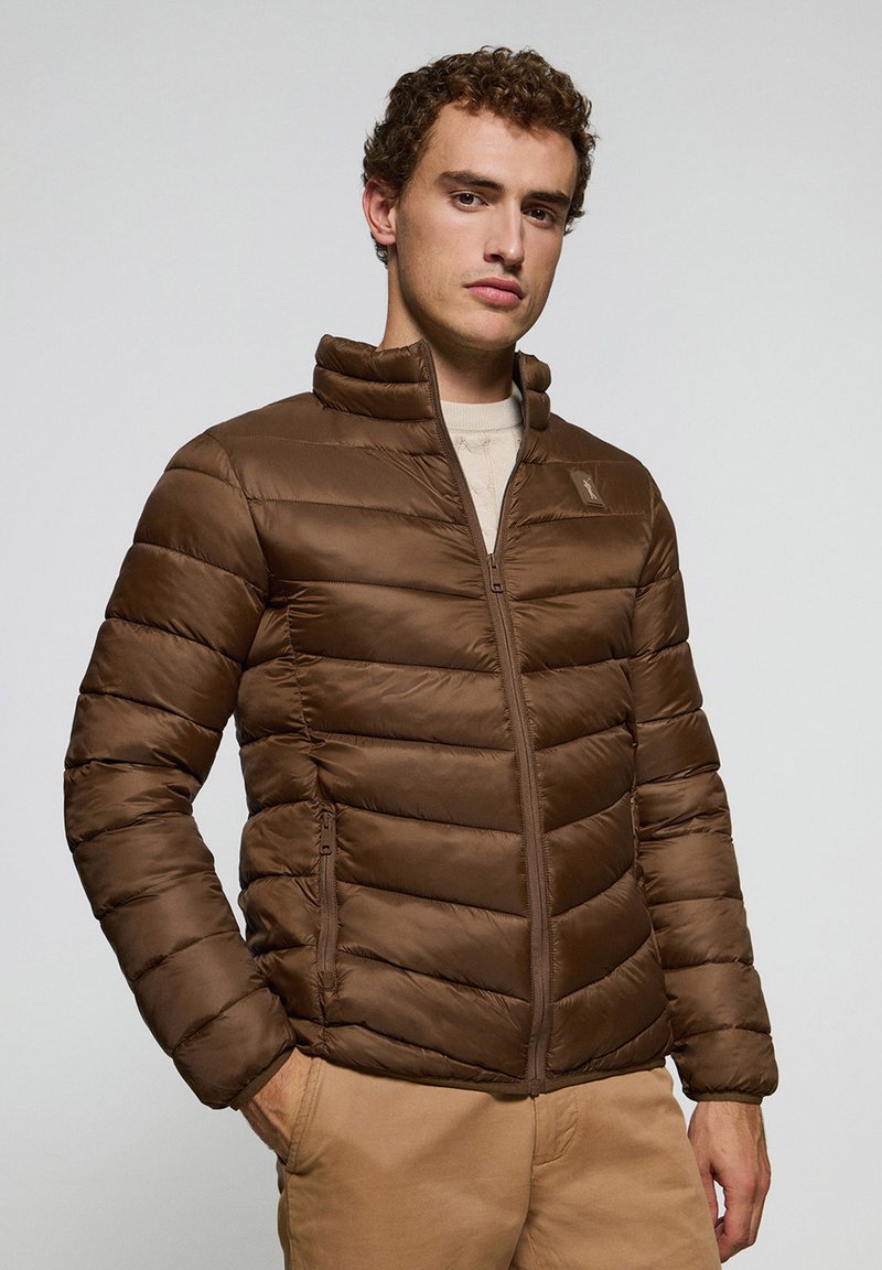 Chaqueta acolchada marrón con diseño de rombos horizontales, cuello alto y cremallera frontal. Presenta bolsillos laterales y una textura suave y brillante.