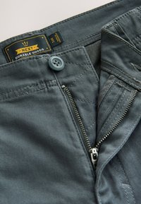 Pantalon gris foncé avec bouton et fermeture éclair partiellement ouverte, avec une étiquette "Next Durable Goods" à l'intérieur de la ceinture.