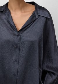 Camicia button-up di un blu navy scuro, realizzata in tessuto stropicciato. Presenta un colletto classico e una vestibilità ampia con bottoni visibili lungo la parte centrale.