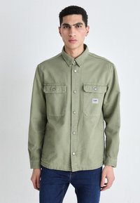 Olive green shirt jacket made of cotton. Features a pointed collar, snap buttons, and two chest pockets. Styled in a relaxed fit.