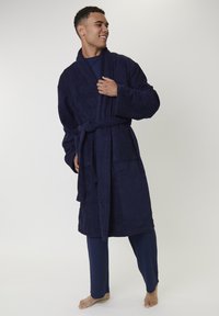 Junger Mann steht barfuß da und trägt einen marineblauen Bademantel mit Taschen, der an der Taille gebunden ist, über einer passenden marineblauen Pyjama-Anlage.