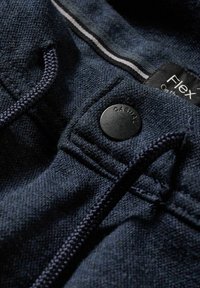 Tela casual de color azul oscuro con un botón negro etiquetado como "CASUAL", un cordón, y una etiqueta de ropa parcial que dice "Flex."