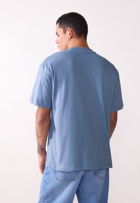 Blå bomulls T-skjorte med korte ermer og rund hals, med en avslappet passform. Brukt med lyseblå denimjeans; enkel bakvisning.