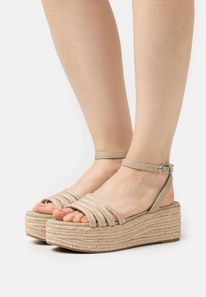 Pieds portant des sandales compensées beige avec des brides tressées et des fermetures à boucle à la cheville sur fond blanc.