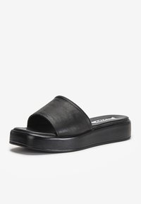Sandale mule en cuir noir avec une large sangle, une semelle plate et un design minimaliste. Présente une texture lisse et un éclat subtil.