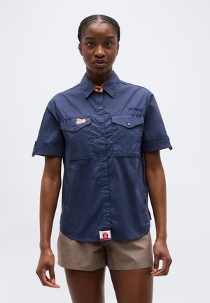 Γυναίκα που φοράει μπλε σκούρο μπλουζάκι με κοντά μανίκια και τσέπες στο στήθος, καθώς και μπεζ σορτς, στέκεται μπροστά σε απλό λευκό φόντο.