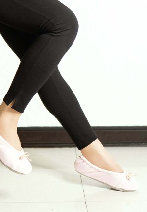 Ballerines rose clair avec une tige en tissu doux, une semelle plate et un nœud décoratif, associées à des leggings noirs ajustés avec des fentes au niveau des chevilles.