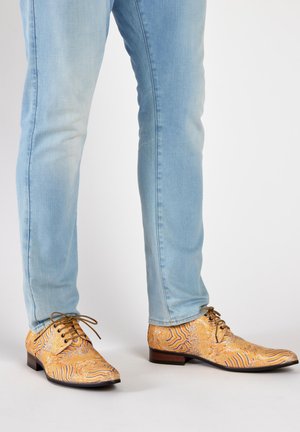 Heren veterschoenen in een intricaat oranje en goudpatroon, met een puntige neus en een bruine hak, gedragen met lichtblauwe jeans.