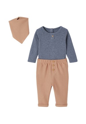 Graues Langarmshirt mit Knopfleiste und einer tanfarbenen Hose aus strukturiertem Stoff mit drei Knöpfen, ergänzt durch ein passendes Bandana.