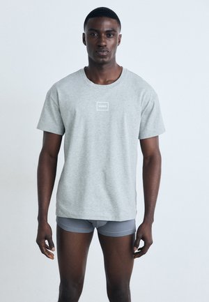 Graues Baumwoll-T-Shirt mit kurzen Ärmeln, mit einem weißen rechteckigen Logo in der Mitte. Trägt man über grauer, taillierter Unterwäsche. Einfarbiger Hintergrund.