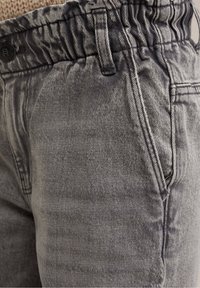 Šedé denimové kalhoty s elastickým pasem, mají přední kapsy a strukturovanou tkaninu. Materiál vypadá vybledle a opotřebovaně.