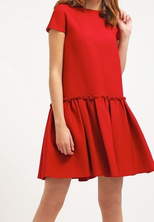 Femme portant une robe rouge à manches courtes, longueur aux genoux, avec une jupe froncée et évasée ainsi qu'un col rond simple.