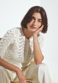 Mujer con cabello corto y castaño, vistiendo un cárdigan blanco de botones tejido y pantalones crema, apoyando la cabeza en su mano, sentada en una silla de mimbre.