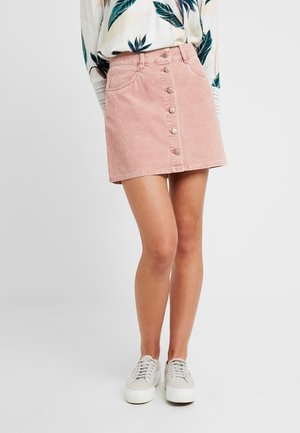 Kvinde iført en pink mininederdel med knapper foran, hvid langærmet bluse med blomsterprint og hvide sneakers står mod en ensfarvet baggrund.