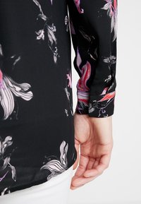 Czarna bluzka w kwiaty z długimi rękawami, w intensywne różowe i fioletowe wzory kwiatowe, z guzikiem na mankiecie rękawa.