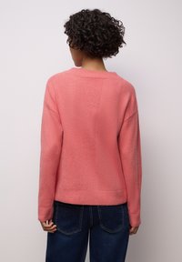 Korallinpunainen neule, jossa on teksturoitu pinta, lasketut olkapäät ja resorit hihansuissa. Yhdistetty tummansinisiin farkkuihin.