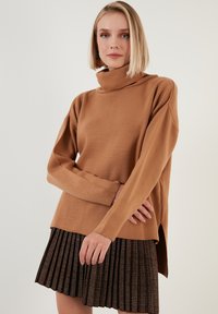 Brauner, langärmeliger Rollkragenpullover mit lockerer Passform und seitlichen Schlitzen, kombiniert mit einem plissierten, strukturierten Rock in Schwarz und Braun.