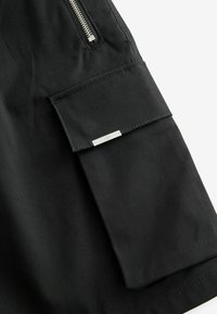Gros plan d'un vêtement en tissu noir présentant une poche zippée et un rabat avec une petite étiquette en métal. Texture lisse avec des détails de couture subtils.