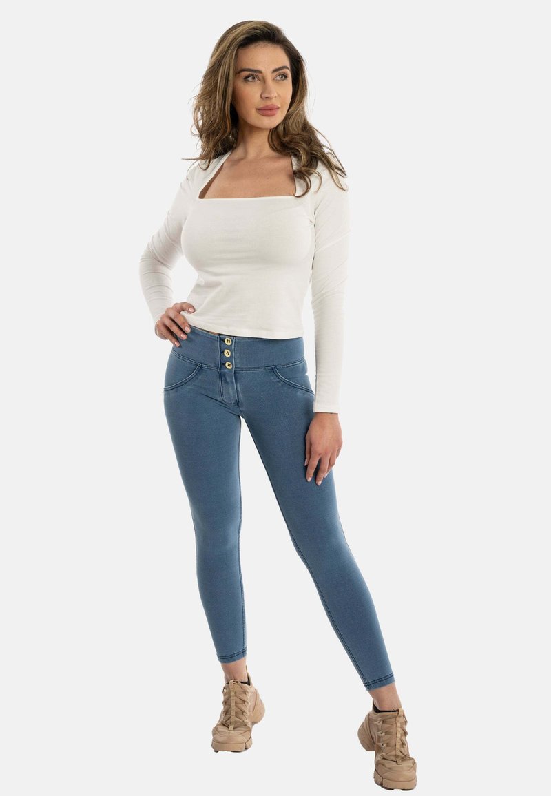 Haut long à manches blanches avec décolleté carré, associé à un jean skinny bleu taille haute avec fermeture à boutons dorés et deux poches latérales.