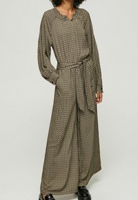 Långärmad jumpsuit i en mörk, mönstrad tyg med V-ringning, bälte i midjan, vida ben och lös passform. Har små geometriska mönster.