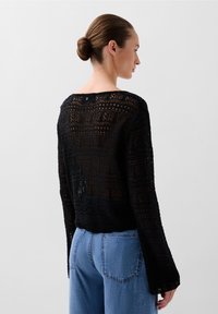 Svart stickad tröja med ett geometriskt spetsmönster, något lös passform, långa ärmar och rundad halsringning. Bärs tillsammans med blå jeans.