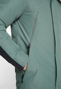 Grön puffjacka med en slät, matt yta, sidoficka med dragkedja och kontrasterande svarta ärmdekorer. Lätt och isolerad design.