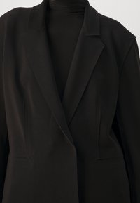 Blazer noir en tissu doux, avec une coupe ajustée, des revers à pointe et des poches avant. Le design est structuré sans fermetures apparentes.