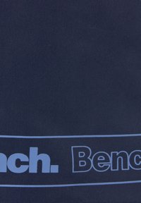 Tkanina tamnoplave boje s grafičkim dizajnom na kojem piše "Bench." svijetloplavim slovima, te s horizontalnom linijom ispod. Glatki, teksturirani materijal.