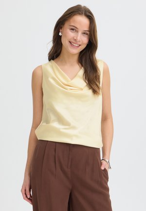 Mosolygó nő hosszú barna hajjal ujjatlan, sápadt sárga, redős felsőt és magas derekú barna nadrágot visel, áll egy kézzel a zsebében.