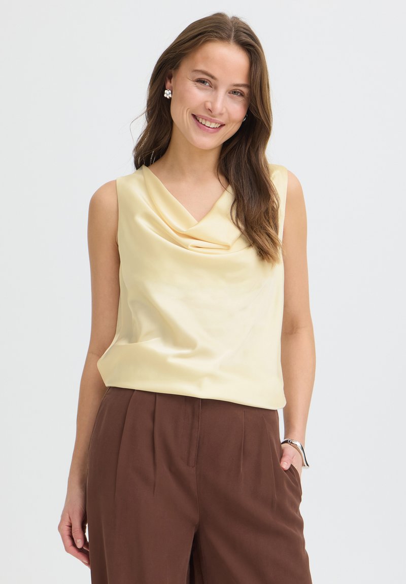 Femme souriante aux longs cheveux bruns porte un top sans manches jaune pâle drapé et un pantalon marron taille haute, debout avec une main dans la poche.
