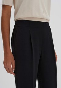 Zwarte, op maat gemaakte broeken met plooien en een elastische tailleband, gecombineerd met een lichte, kortgeïnen crèmekleurige top. Glad textiel.