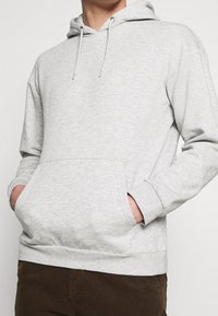 Sudadera gris claro con capucha, cordones, bolsillo delantero y puños acanalados. Textura de tela suave con un corte relajado.