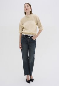 Beige kötött pulóver magas nyakkal, sötét mosású, egyenes szabású farmernadrággal, amelynek szegélyénél foszlott a szövet, és fekete, hegyes orrú magassarkú cipővel.