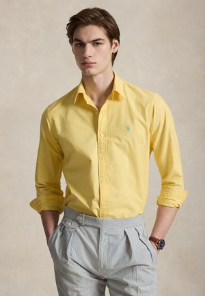 Fiatal férfi barna hajjal, aki sárga, felgyűrt ujjú ingben, csíkos, magas derekú nadrágban, barna órával pózol semleges háttér előtt.